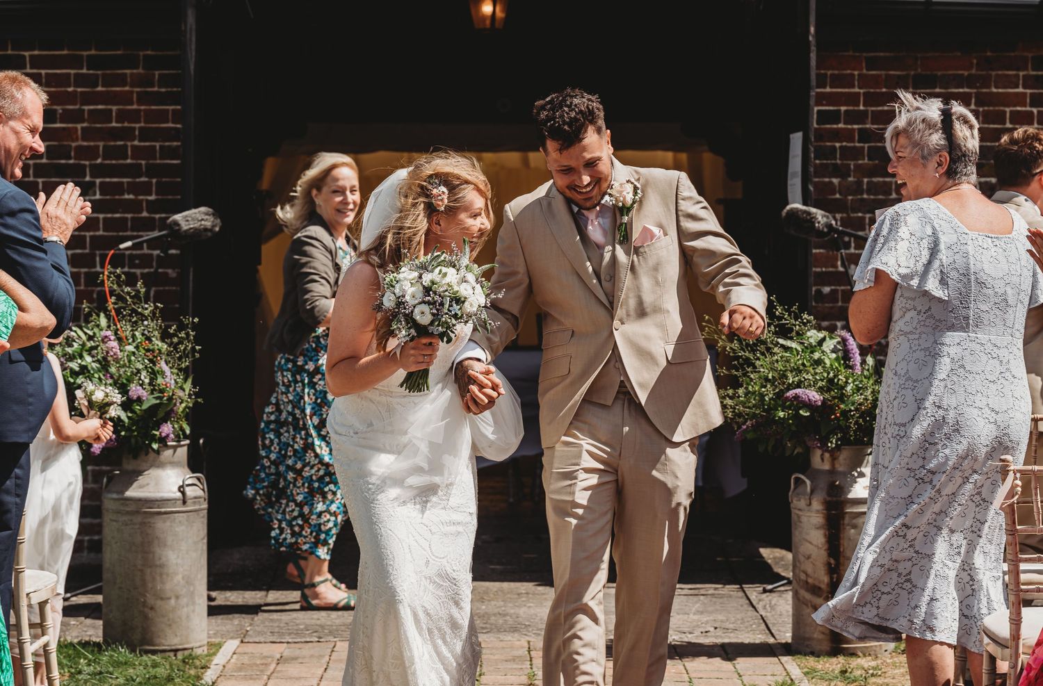 Happy couple exits wedding ceremony in tan suit and white lace dress as guests throw flower petals outside brick venue.