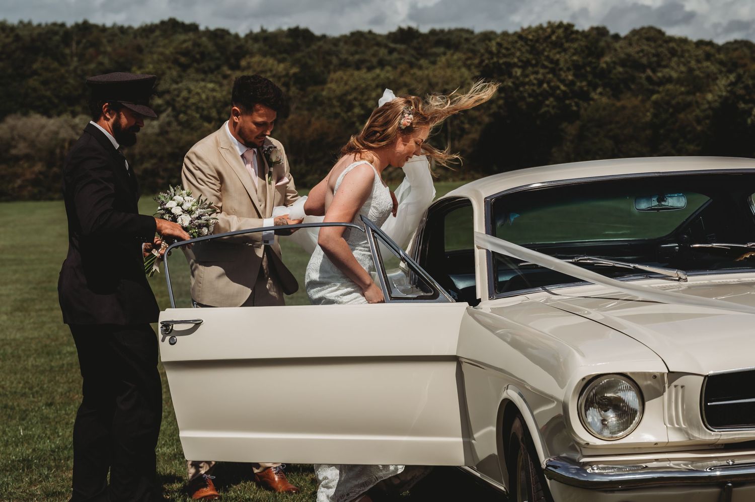 Wedding party gathered by a classic white Ford Mustang on a green countryside field under cloudy skies.