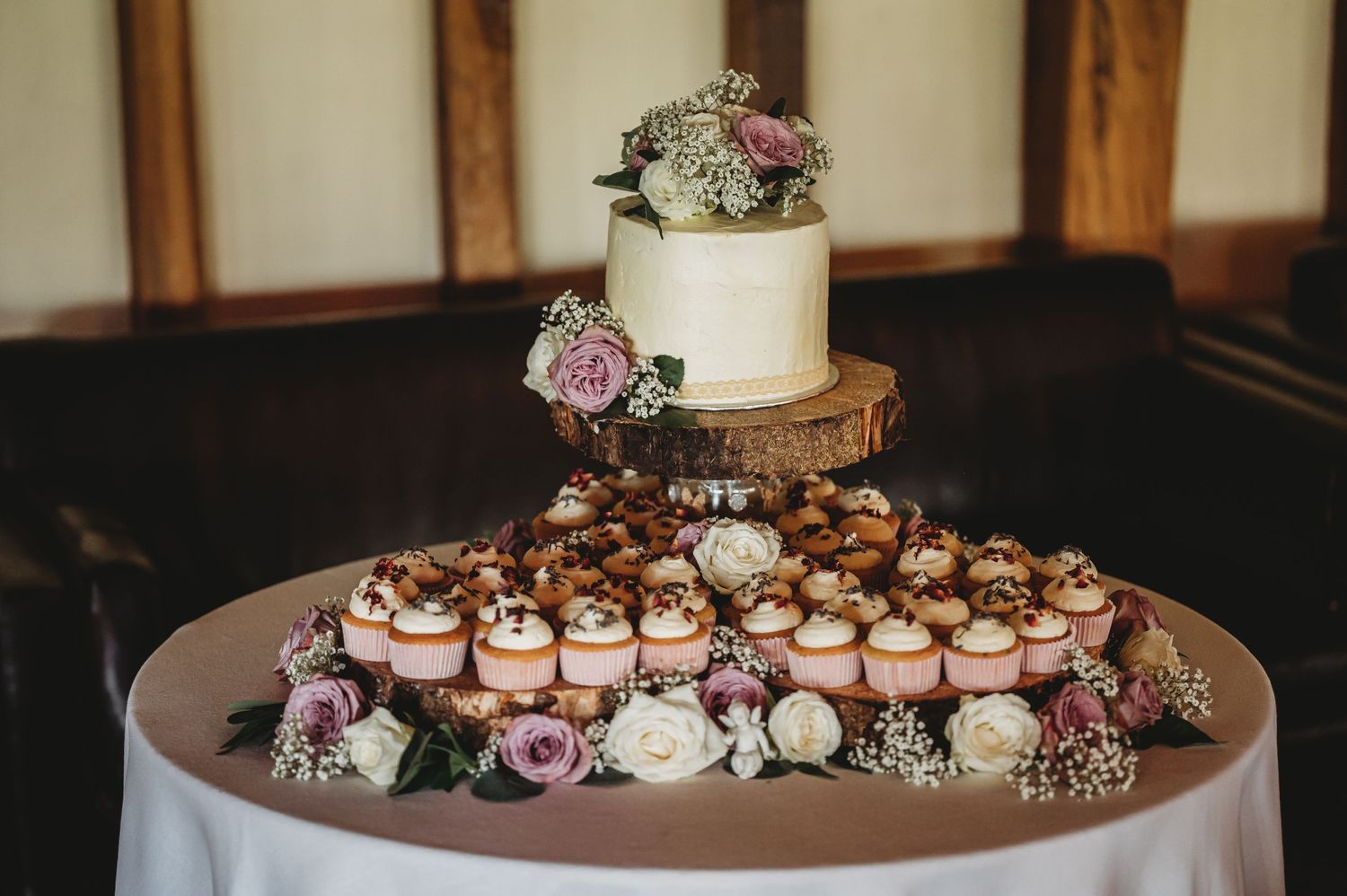 A wedding dessert display featuring a small white cake and pink cupcakes arranged on a round table with floral decorations.