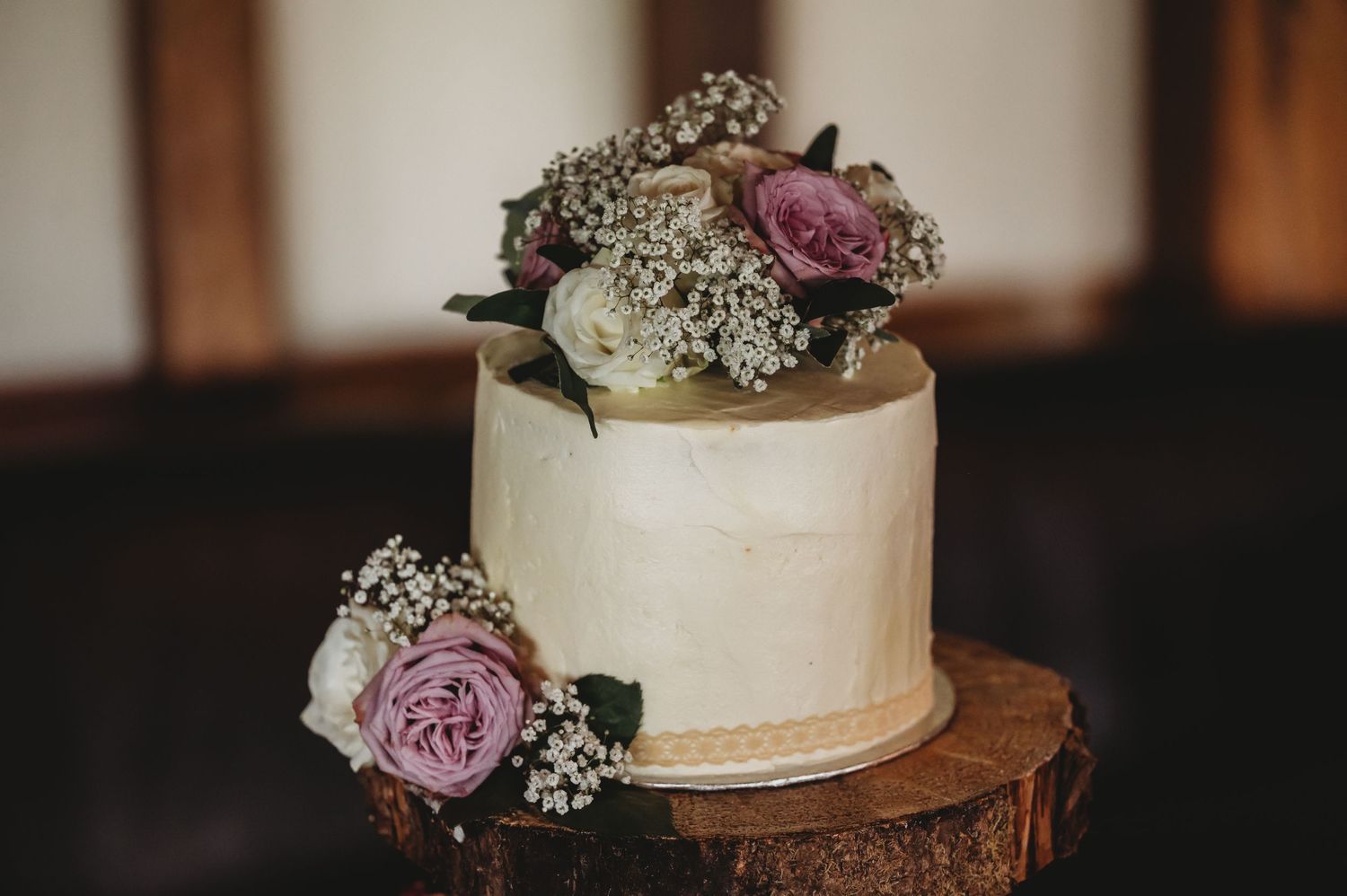 Simple white buttercream wedding cake decorated with mauve roses and baby's breath sits on rustic wooden stand.
