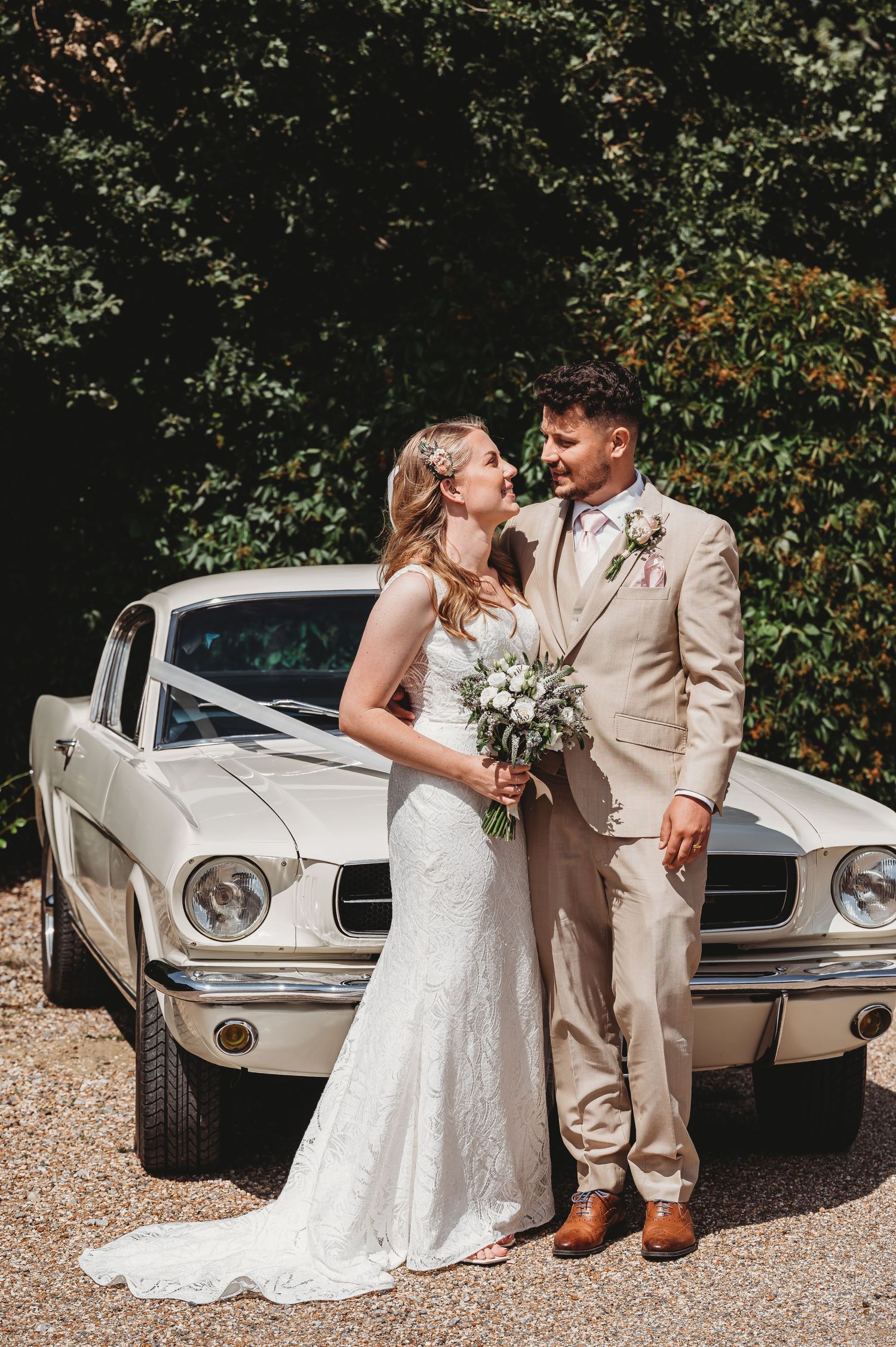 Newlyweds share romantic moment in front of classic white Ford Mustang car during outdoor wedding photoshoot.