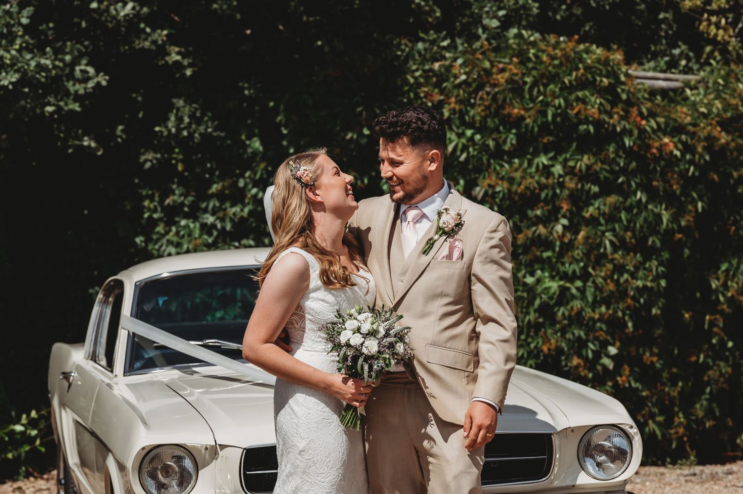 A romantic wedding couple poses together in front of a classic white vintage car on their special day.
