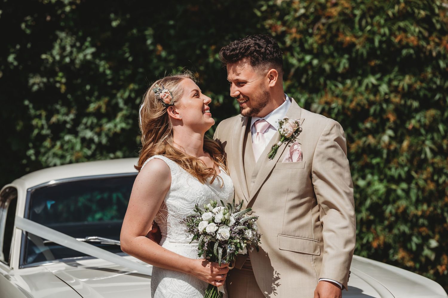 A bride and groom share an intimate moment beside a vintage car while holding a white floral bouquet.