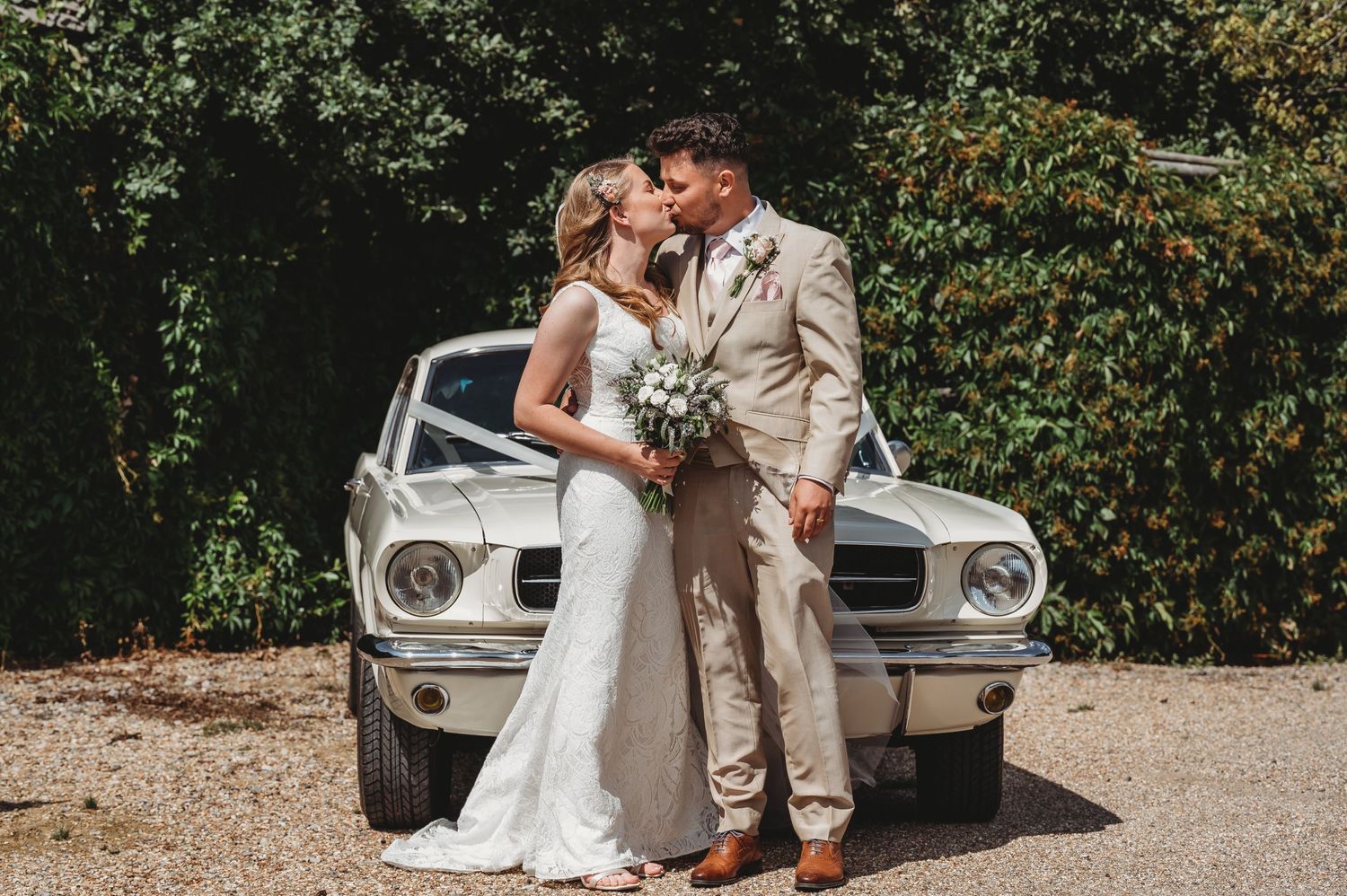 A couple in wedding attire pose romantically in front of a classic vintage white Ford Mustang.