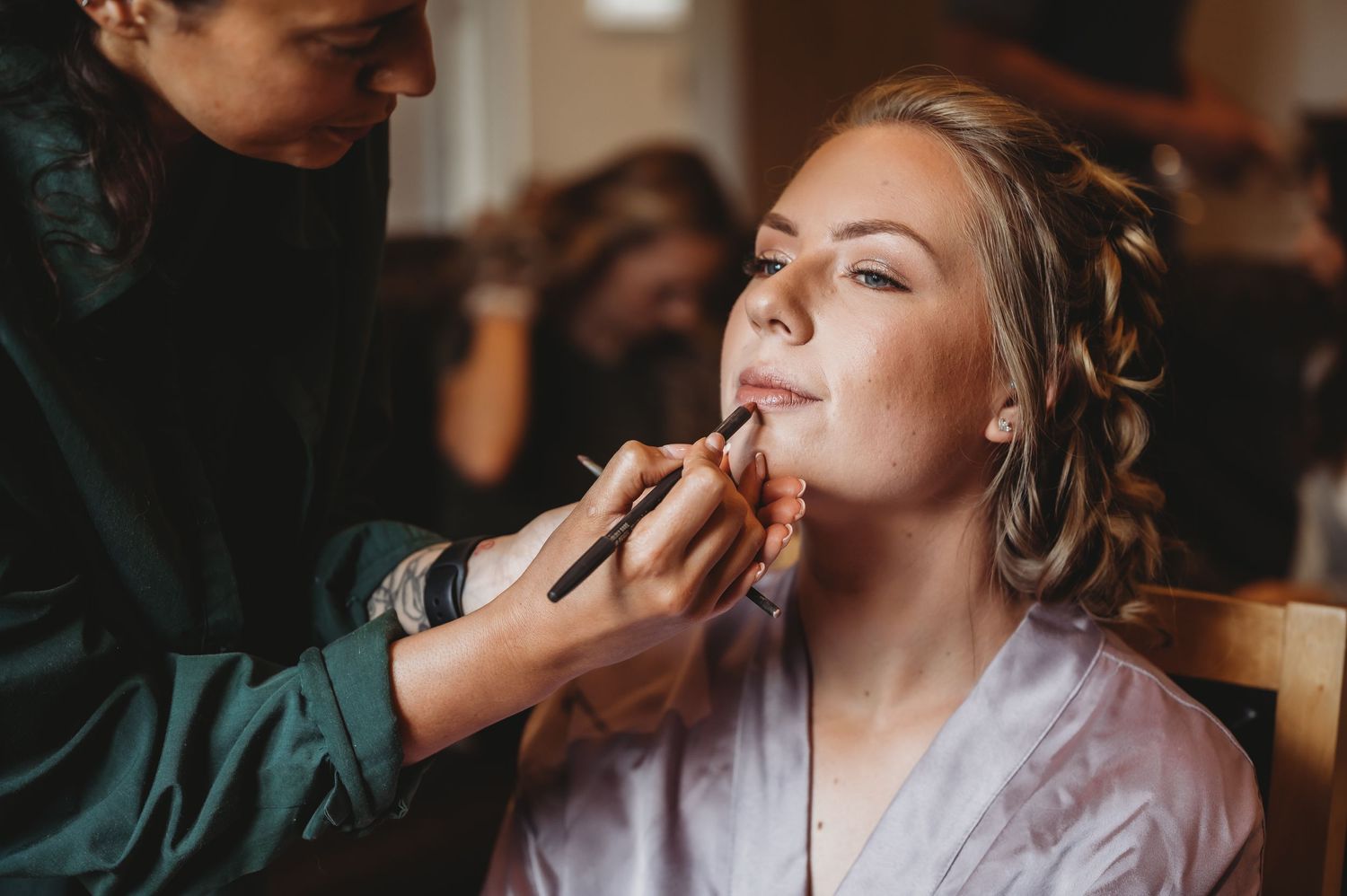 A makeup artist applies lipstick to someone wearing a silk robe during wedding preparations.