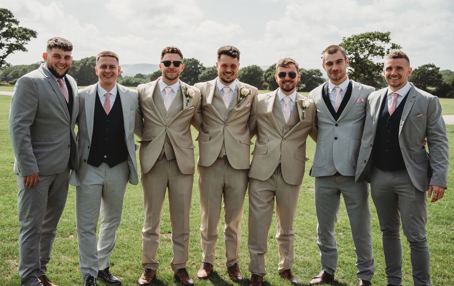 Groomsmen in coordinated light gray and tan suits pose together on grassy field at outdoor wedding.