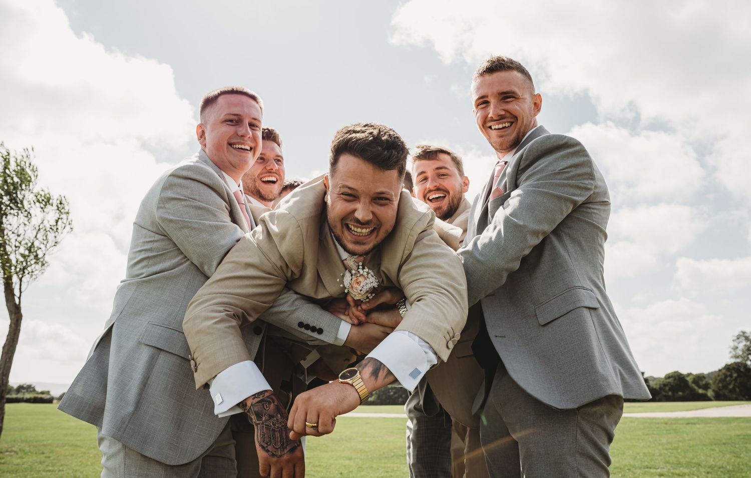 A group of groomsmen in light gray suits playfully pose together outdoors on a cloudy day.