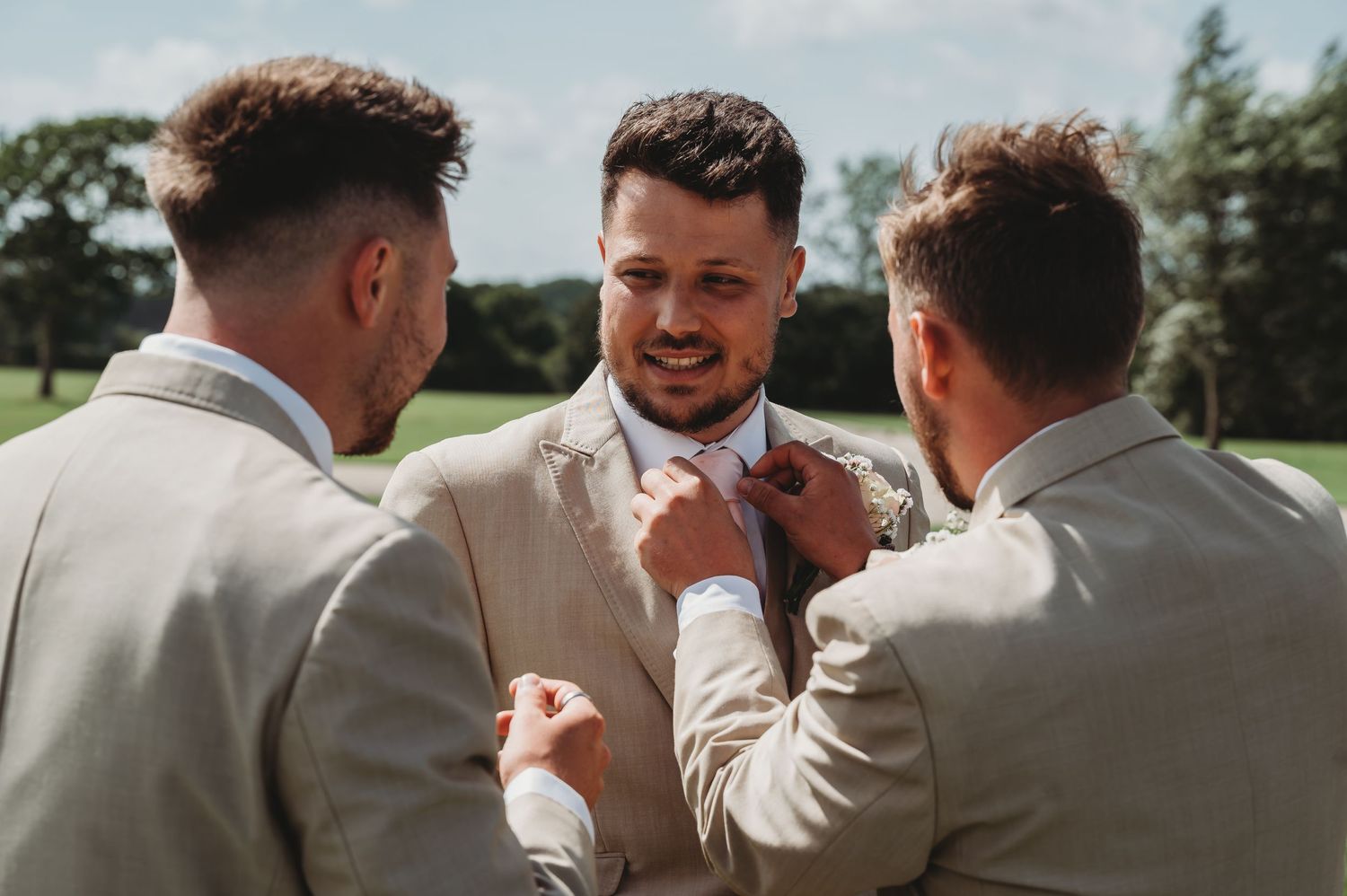 Two groomsmen help adjust a white bowtie on a groom's collar during an outdoor wedding preparation.