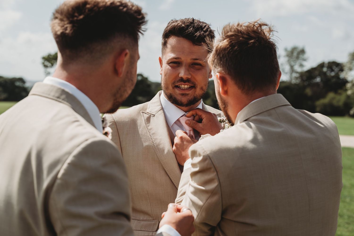 Three groomsmen in light colored suits share a conversation during an outdoor wedding celebration.