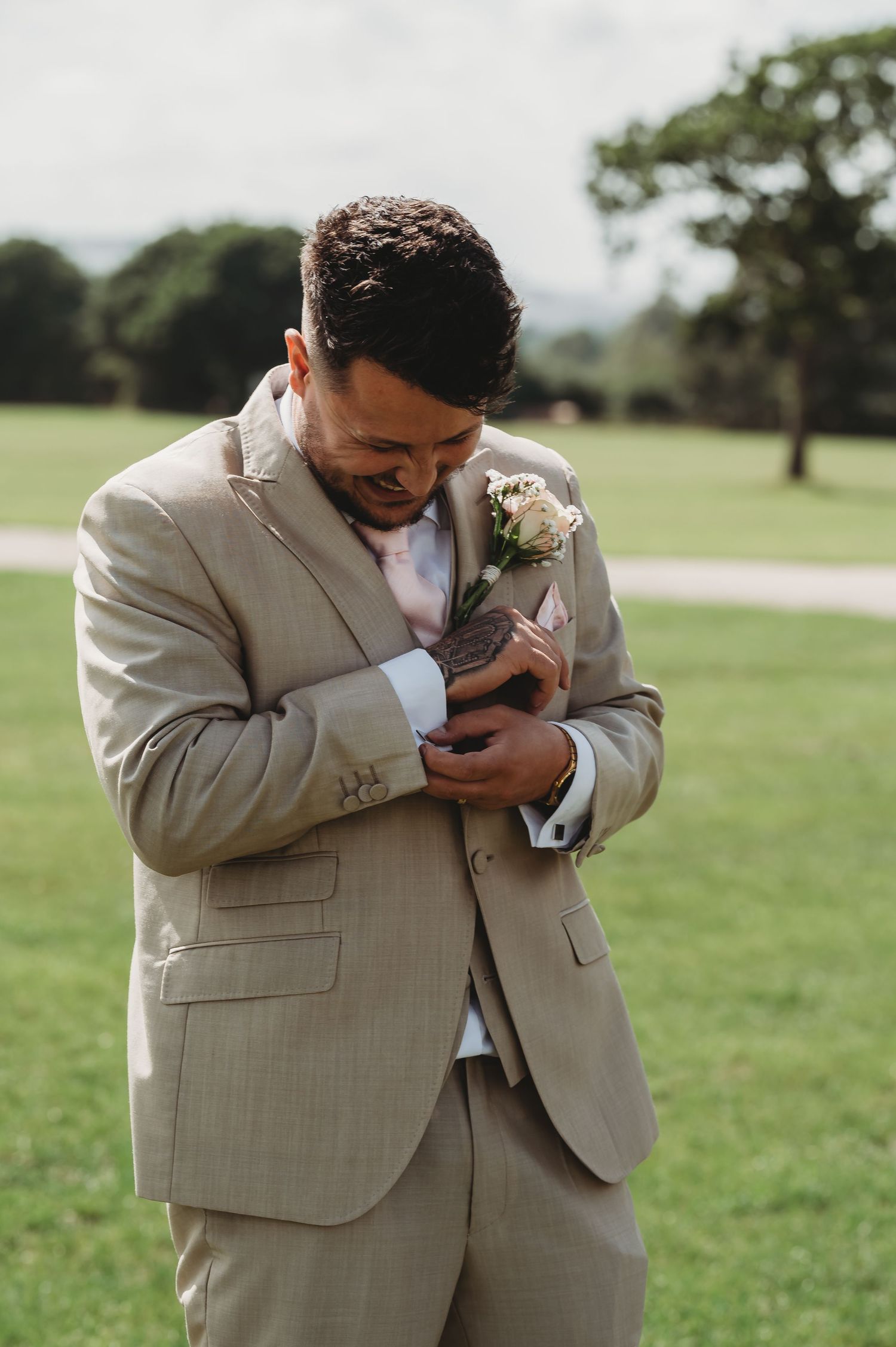 An emotional moment outdoors as groom in beige suit wipes tears during a wedding ceremony on a grassy lawn.