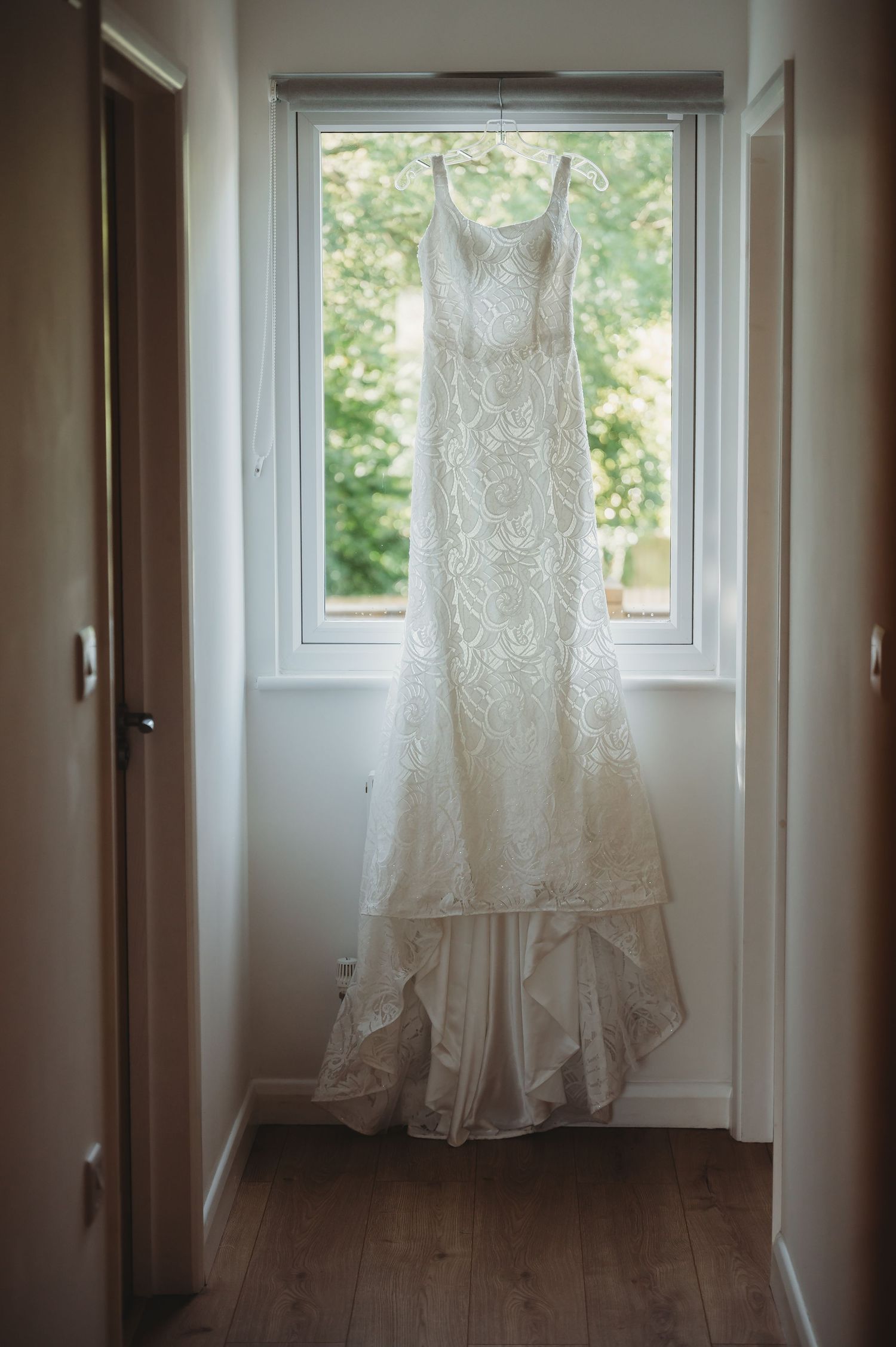 A white lace wedding gown with a mermaid silhouette hanging in front of a bright window.