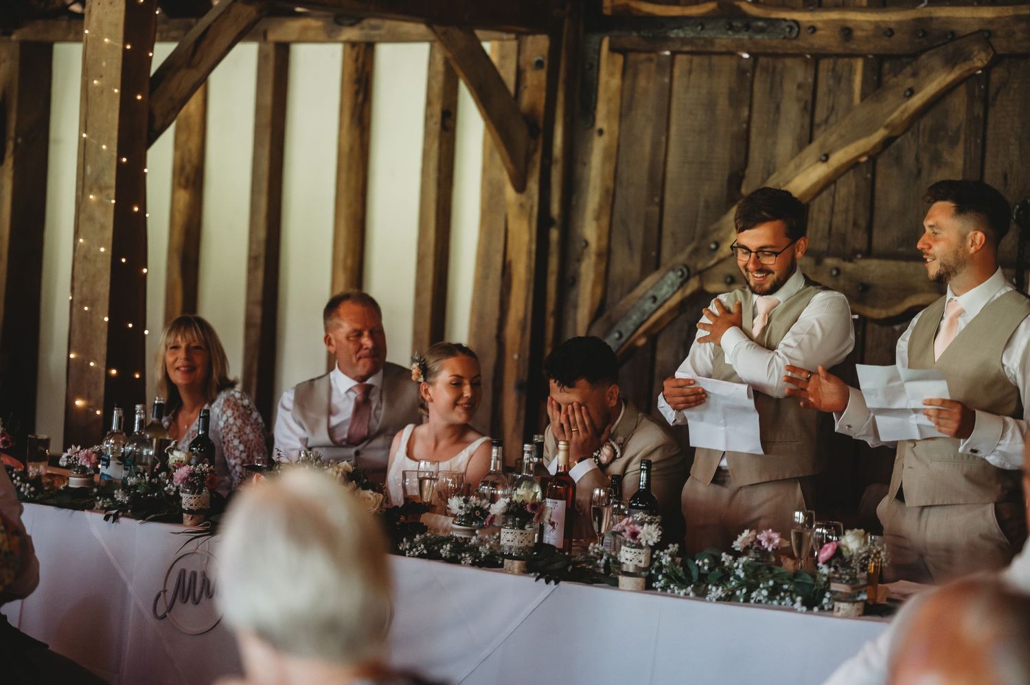 Wedding party seated at head table in rustic barn while speakers give toasts at elegant country reception.