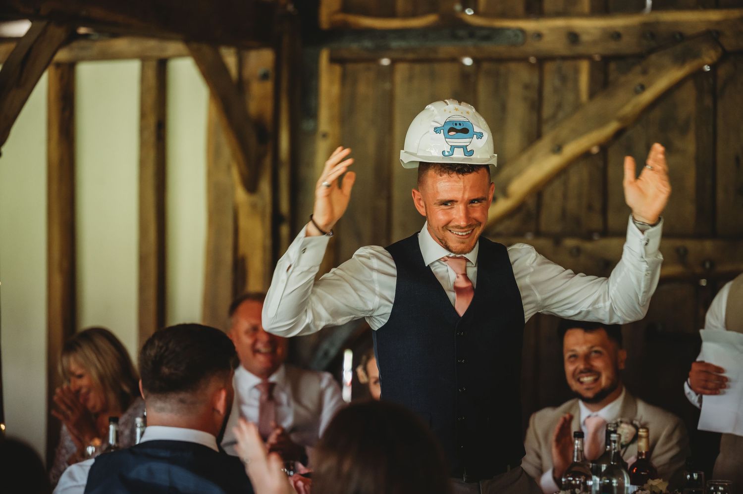 A person in a hard hat and vest raises their hands in celebration at a rustic indoor venue during a wedding reception.
