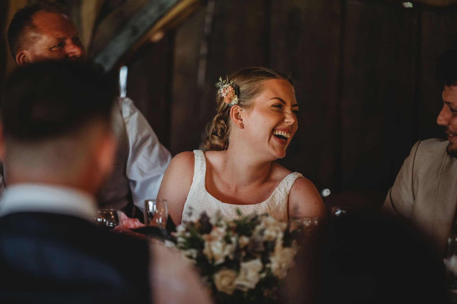 A joyful bride laughs heartily during her wedding reception while seated next to floral table arrangements.