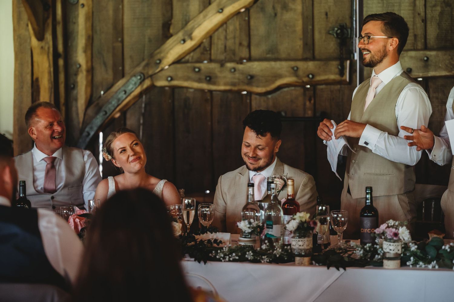 A joyful wedding reception scene with people seated at a dining table while someone delivers a speech in a rustic barn setting.