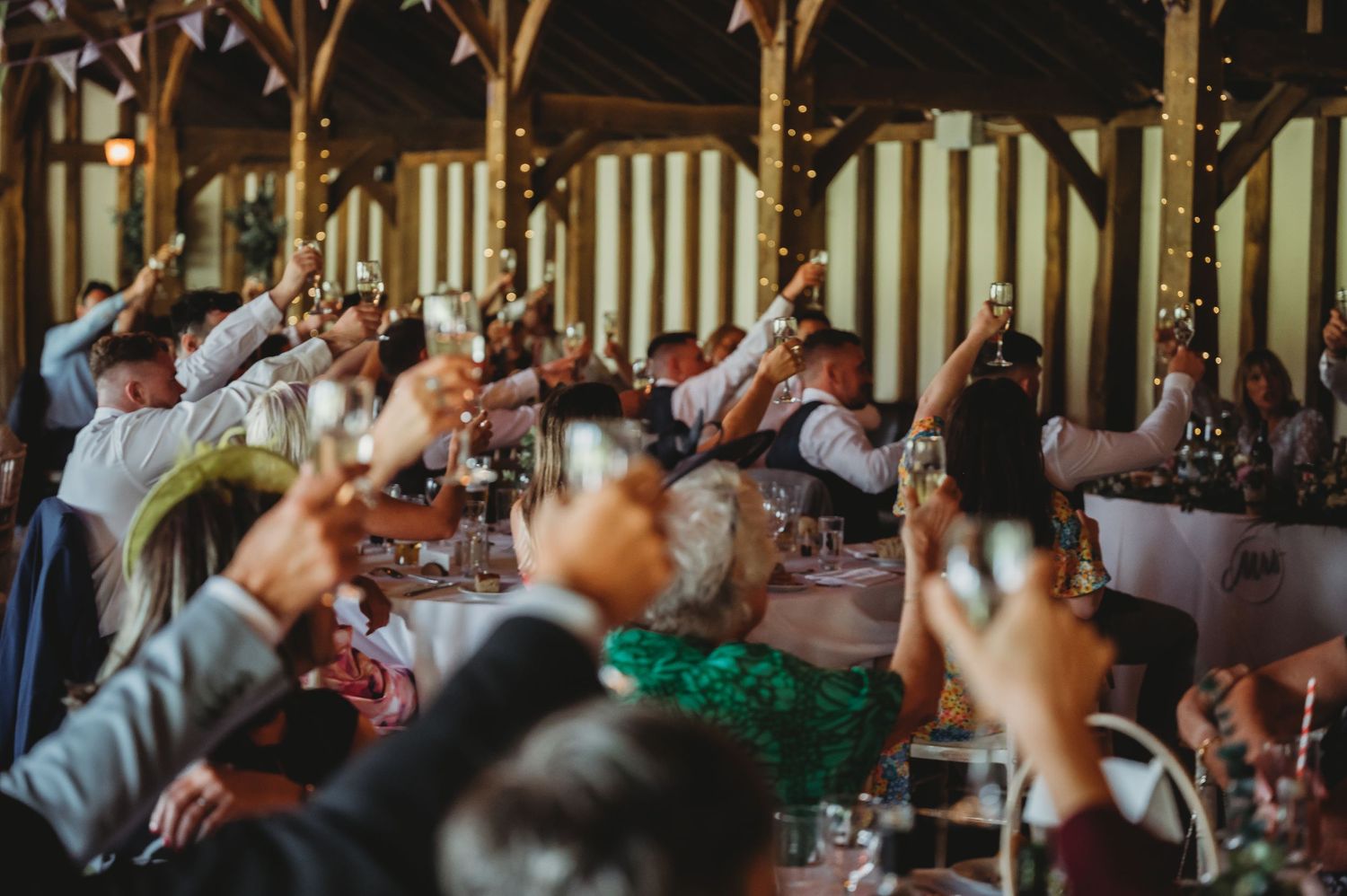 Guests raise their glasses in a toast during a celebration in a rustic wooden venue decorated with overhead beams.