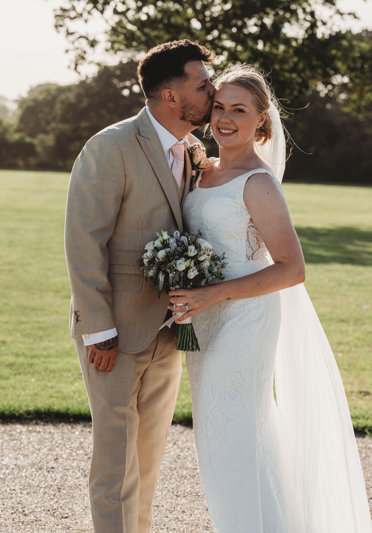 Newlywed couple shares intimate moment outdoors in sunlight with bride holding white floral bouquet.