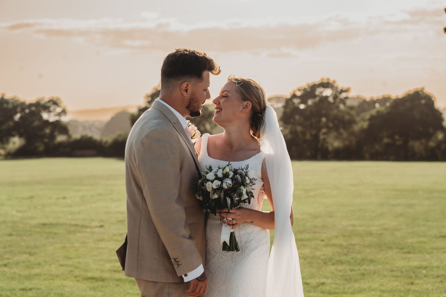 A romantic wedding couple embrace in a sun-drenched field during golden hour with a floral bouquet.