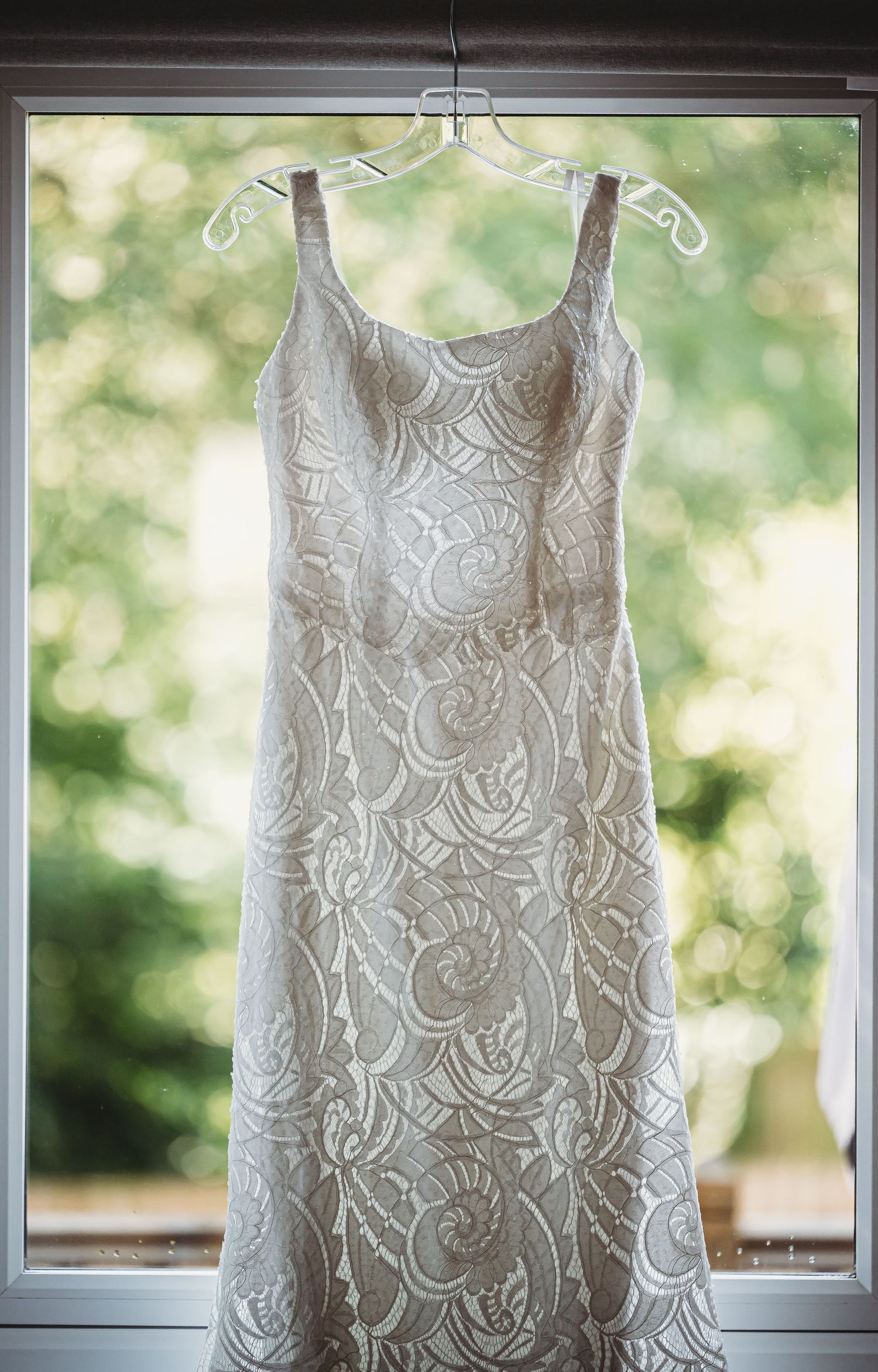White lace wedding dress hangs delicately from a hanger in front of a window with soft natural lighting.