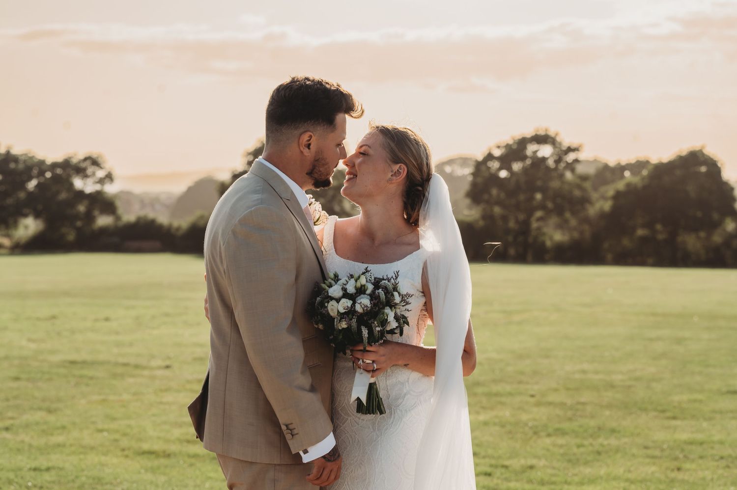 A romantic wedding photo of newlyweds sharing a kiss in a sunlit field while holding a bouquet of white flowers.