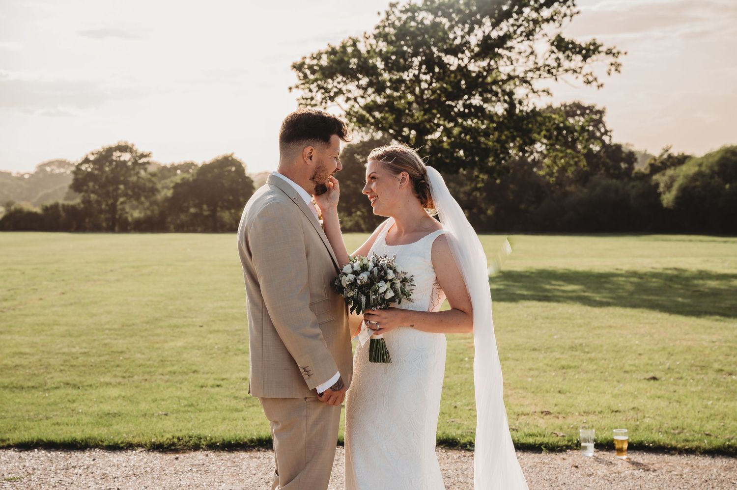 A couple in wedding attire stands together in a sunlit field holding hands during a romantic golden hour moment.