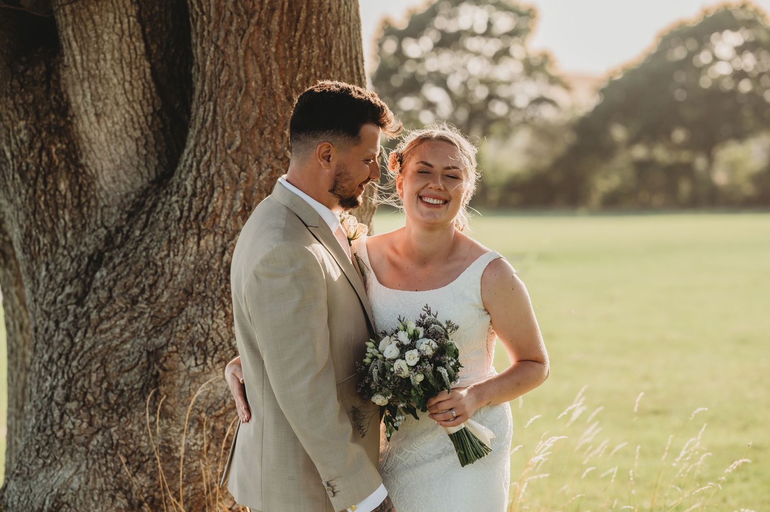 Newlywed couple shares intimate moment by large tree trunk during golden hour sunset in outdoor wedding setting.