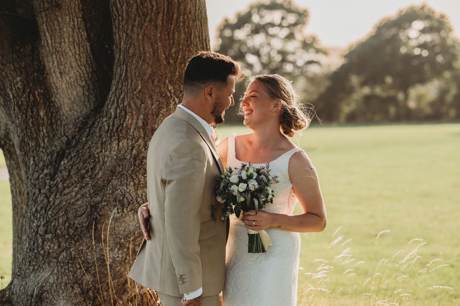 A romantic wedding couple shares an intimate moment together under a large tree in golden afternoon sunlight.