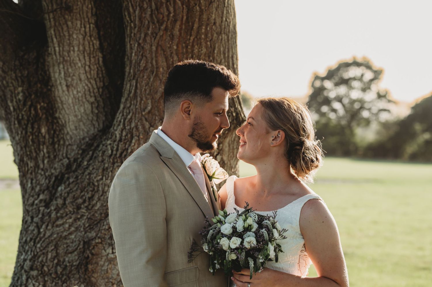 A romantic wedding couple shares an intimate moment beneath a large tree during golden hour, holding a white floral bouquet.