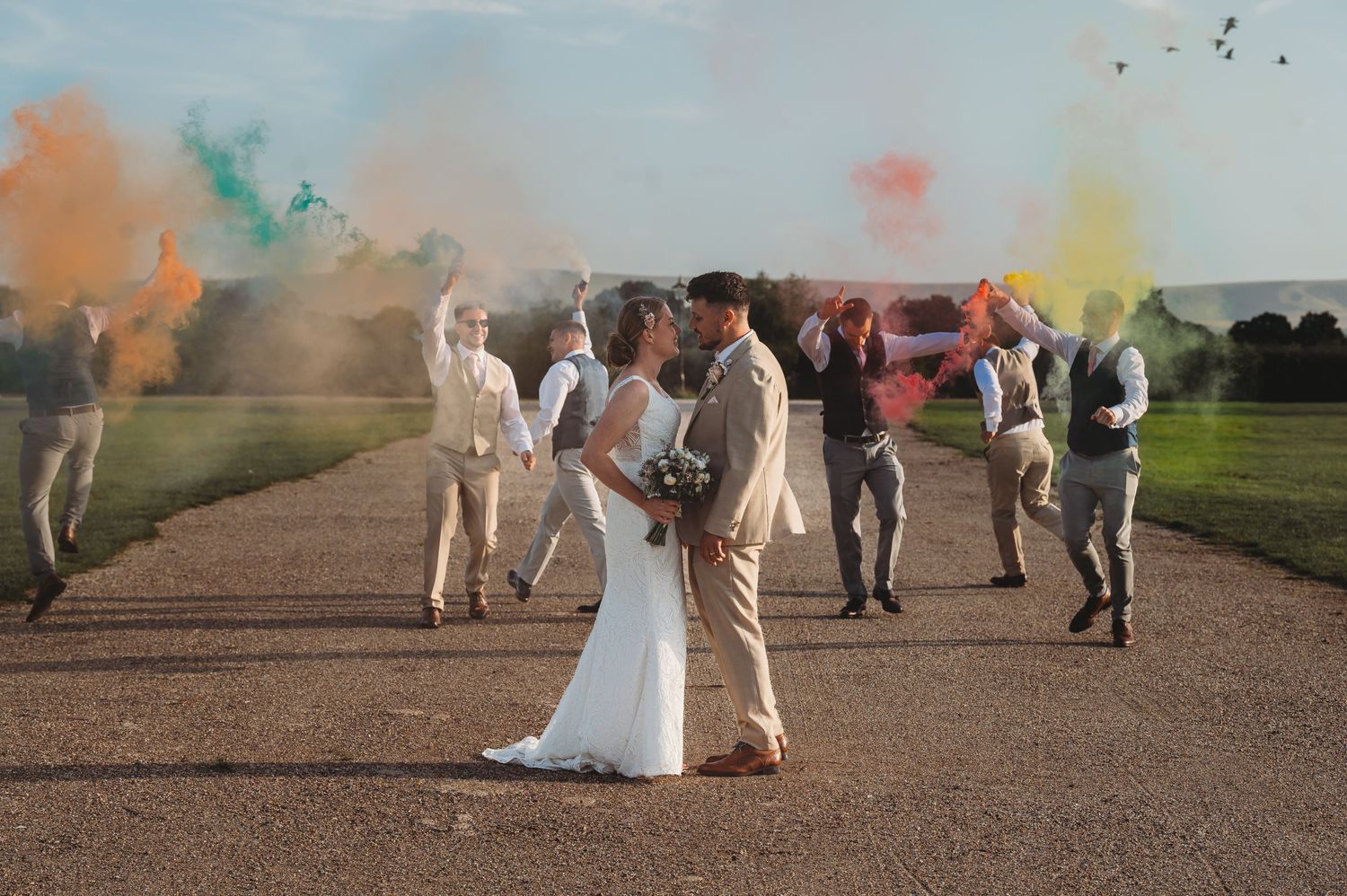 A wedding couple embraces on a path while guests celebrate with colorful smoke bombs in the background.