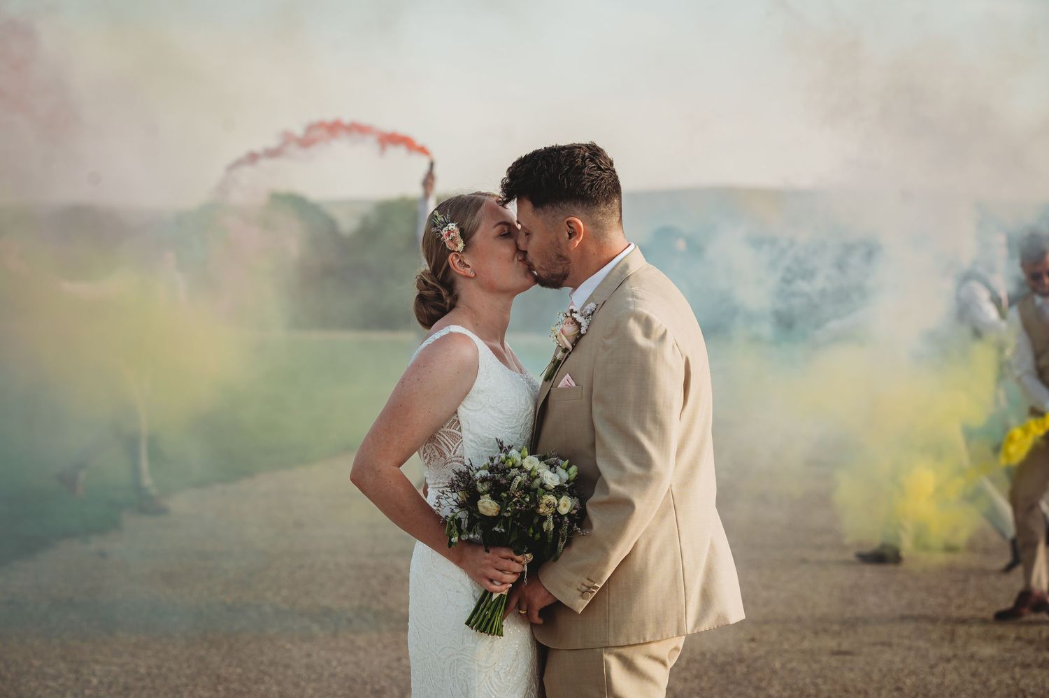 A couple shares a romantic kiss surrounded by colorful smoke during their outdoor wedding ceremony.
