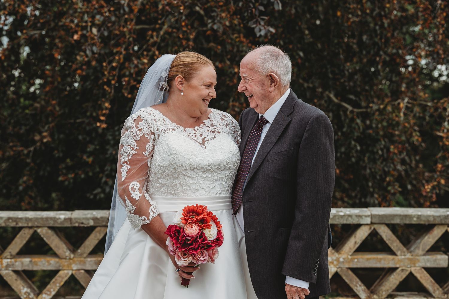 A bride in a lace wedding dress holds a red bouquet while sharing a moment with a person in a dark suit outdoors.