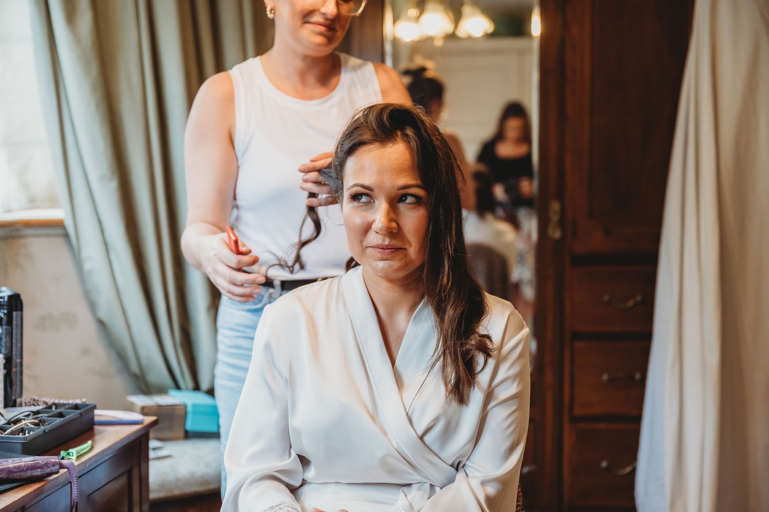 Someone in a white robe sits at a vanity while getting their hair styled in a sunlit room.