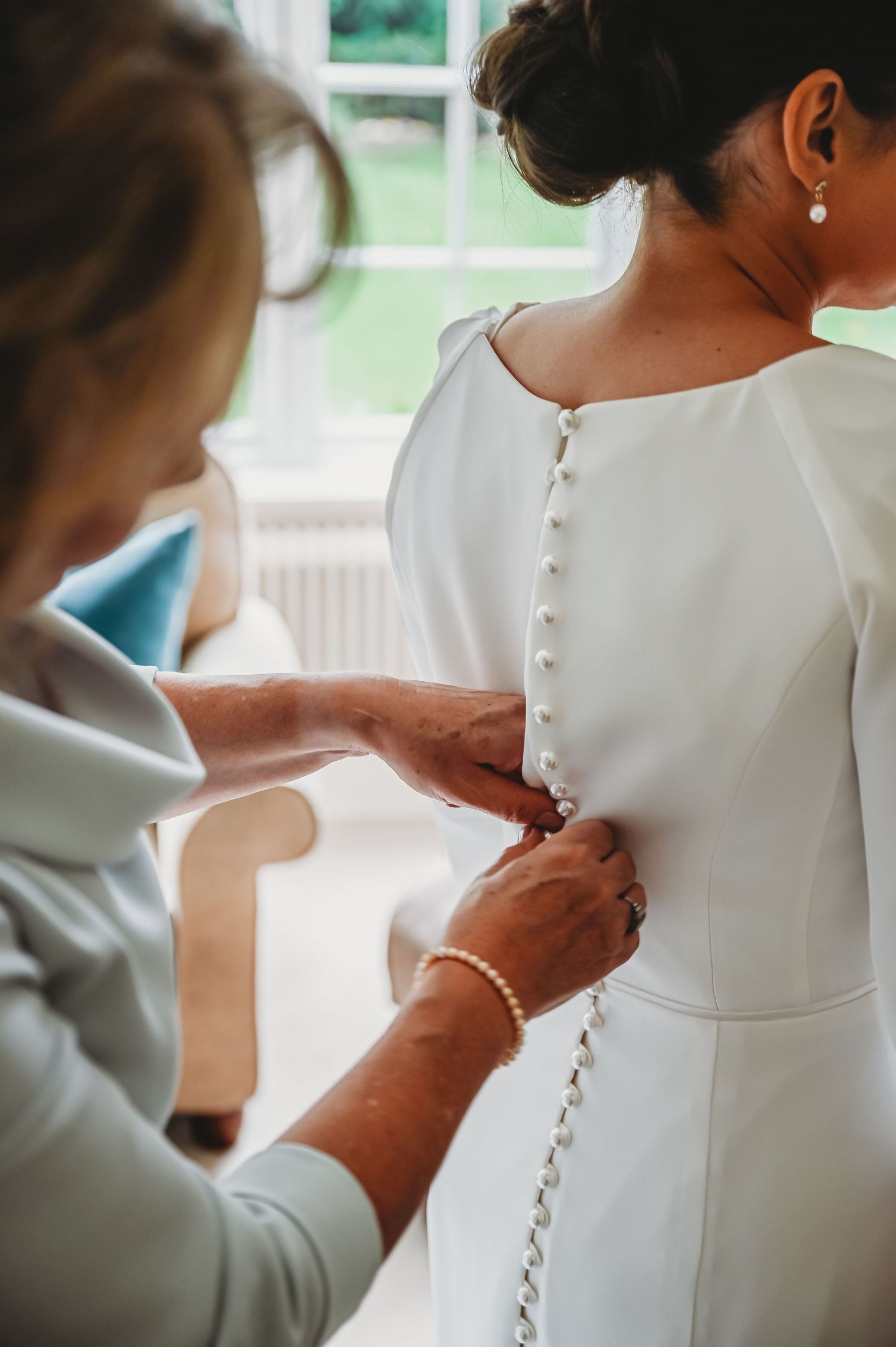 Hands carefully fastening delicate pearl buttons on a classic white wedding dress in a bridal suite.