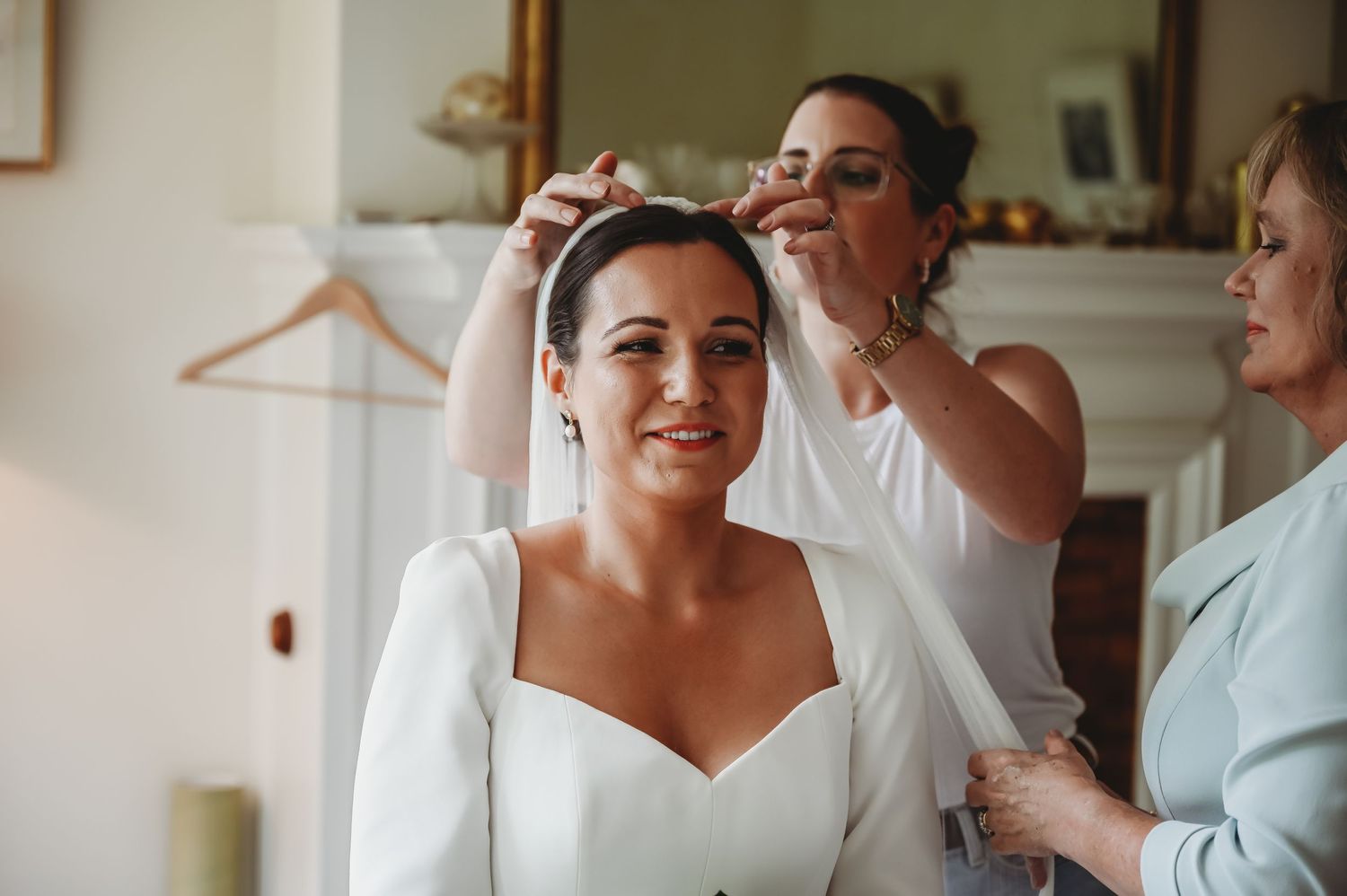 A bride in a white sweetheart neckline dress smiles while getting ready with help from others.