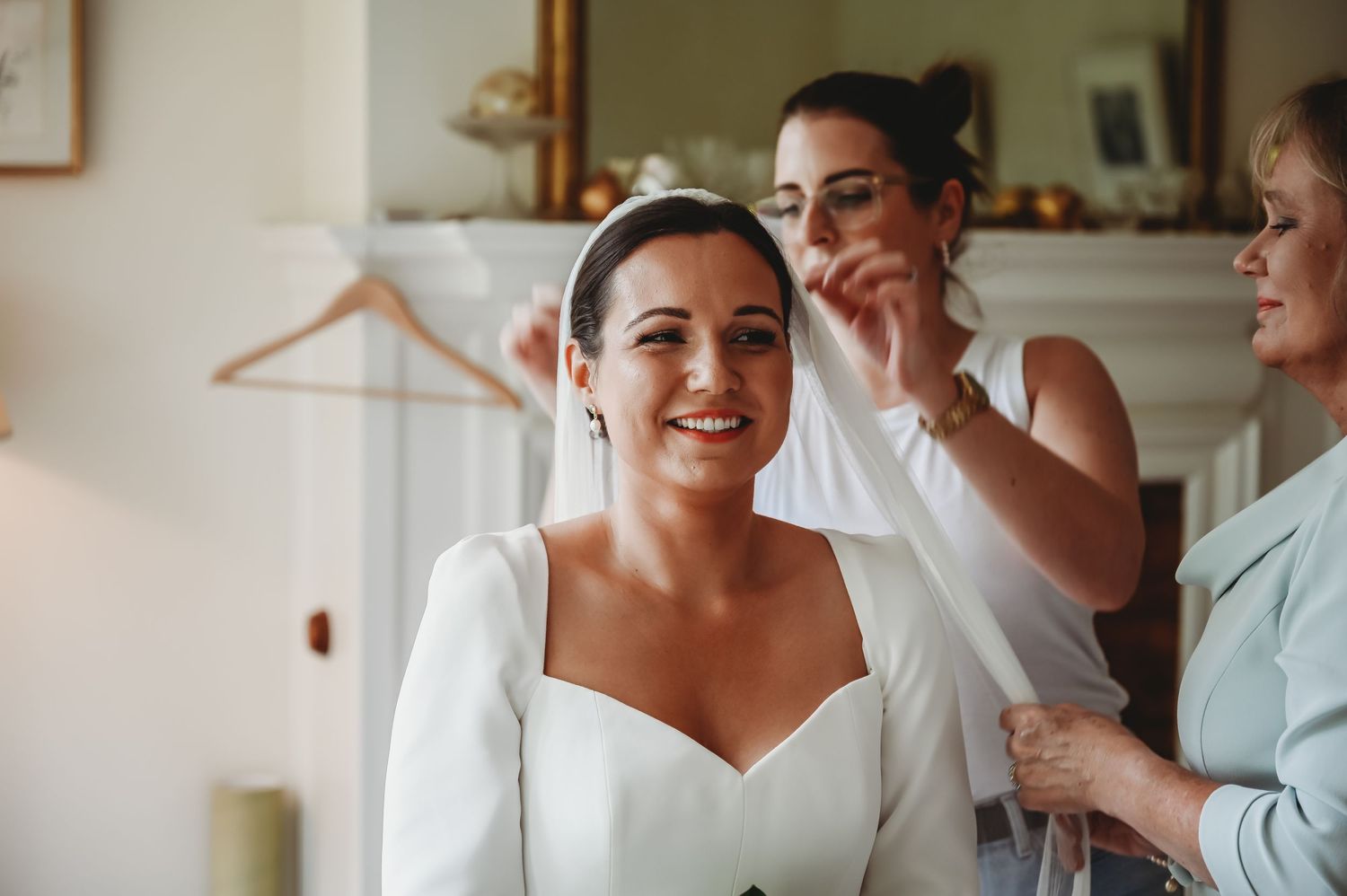 Someone in a white wedding dress smiles while getting their hair styled during wedding preparations.