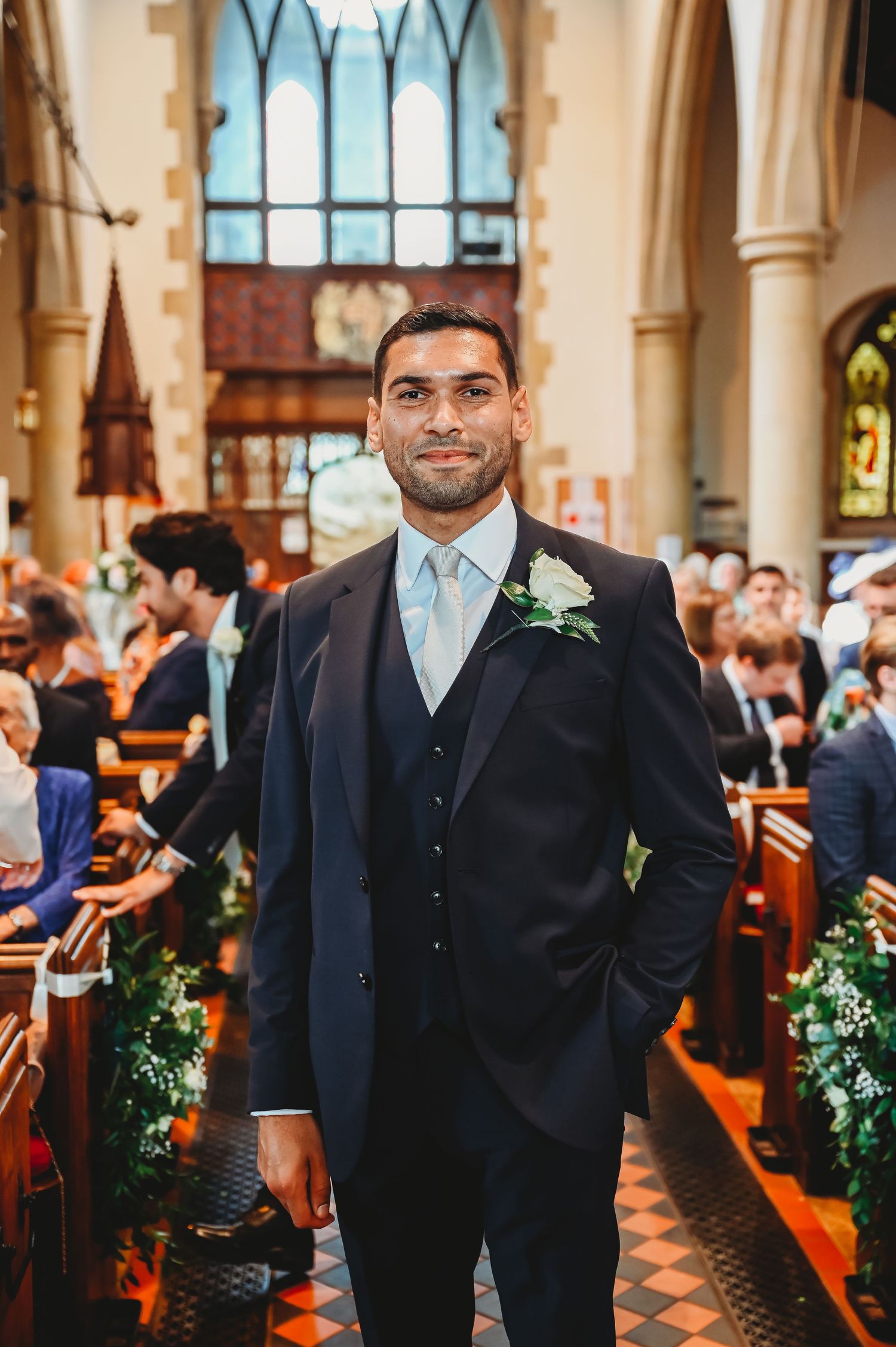 A well-dressed groom in a dark suit with light blue tie stands in the aisle of a church during a wedding ceremony.