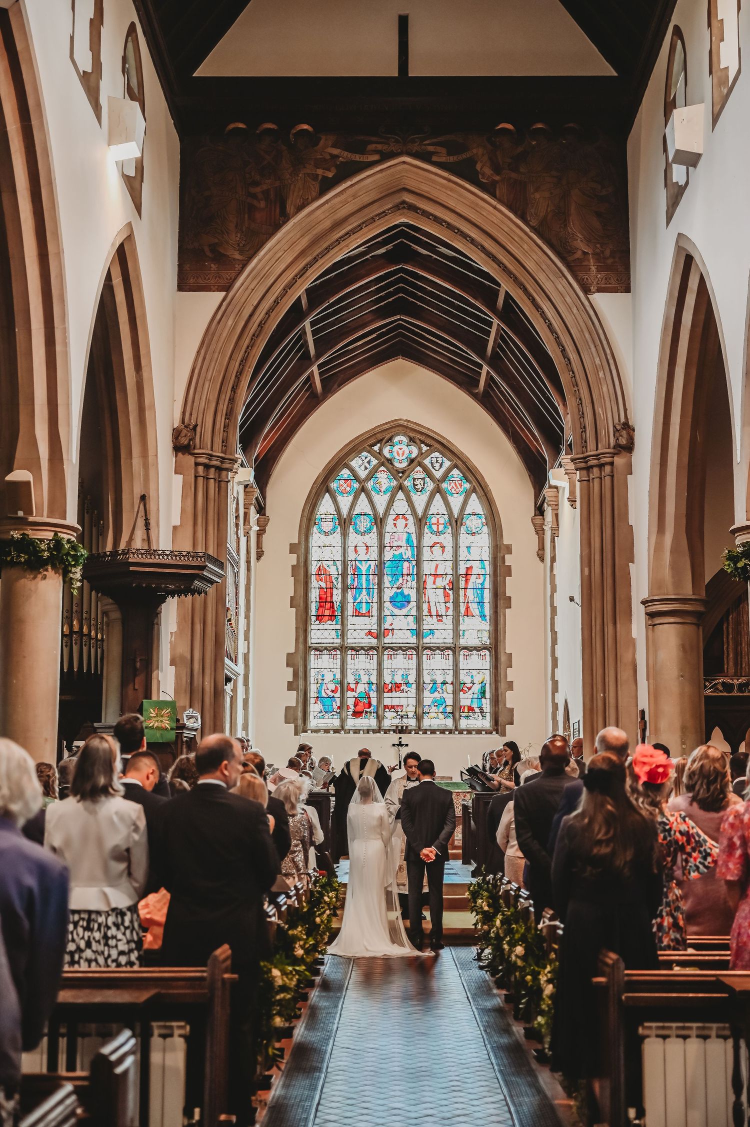 A traditional church wedding ceremony with guests seated in pews beneath Gothic arches and stained glass windows.