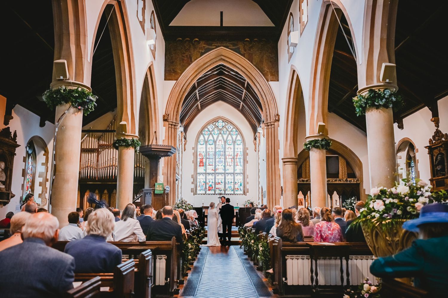 A wedding ceremony takes place in a traditional English church with Gothic arches and stained glass windows.