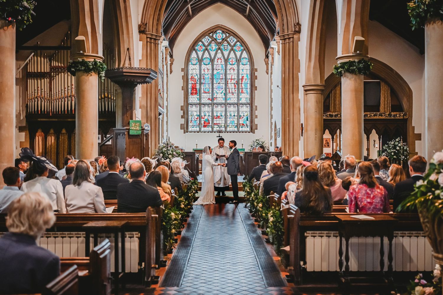 View down church aisle during wedding ceremony with guests seated in wooden pews beneath arched stained glass window.