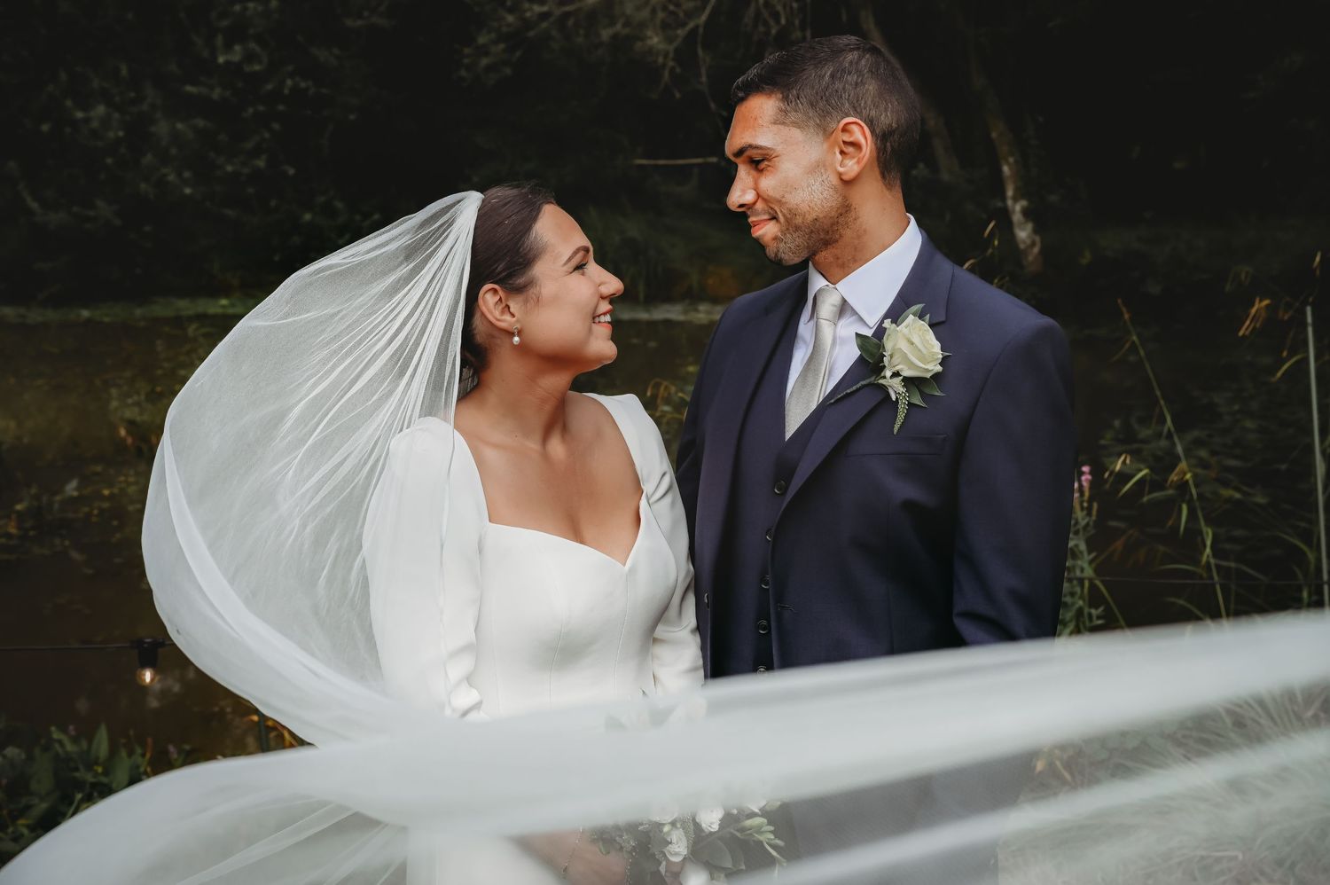 A bride and groom share an intimate moment as her veil flows in the wind against a dark natural backdrop.