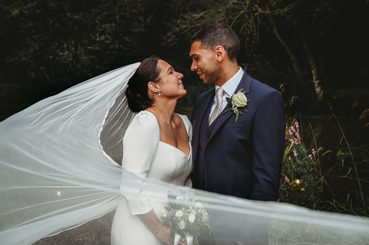 A bride in a white long-sleeved gown and groom in a navy suit share an intimate moment with a flowing veil in nature.