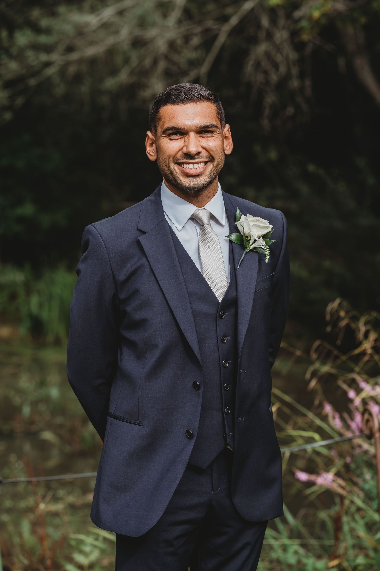 A smiling groom in a dark blue three-piece suit with a grey tie and boutonniere stands outdoors.