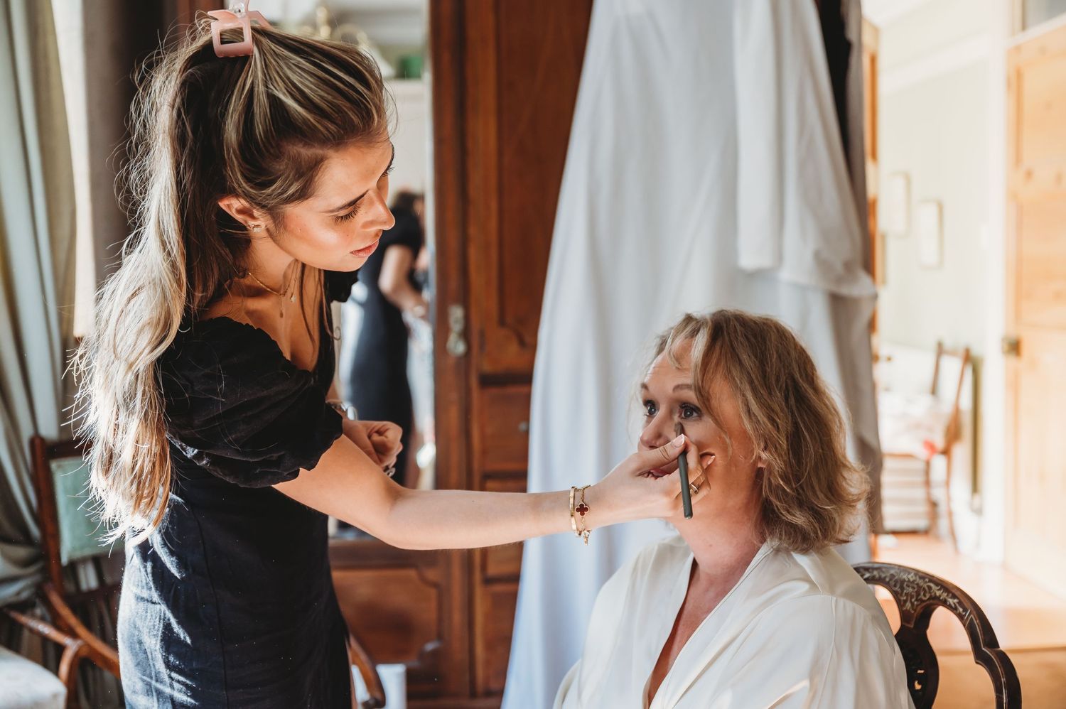 Makeup artist applies cosmetics to someone wearing a white robe in a warmly lit room.