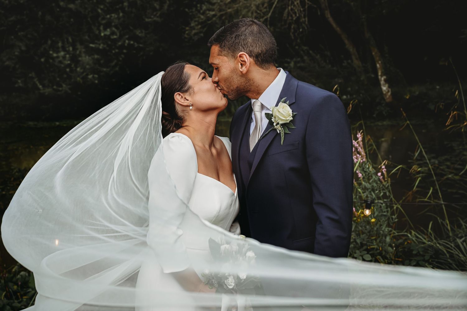 A romantic wedding couple shares a kiss outdoors as the bride's flowing white veil catches the breeze.