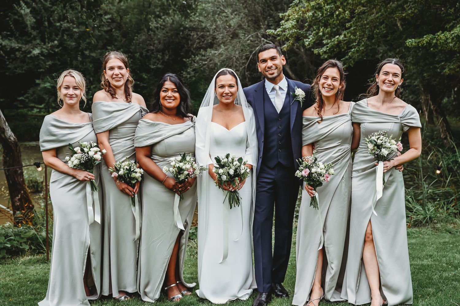 Wedding party wearing elegant light gray gowns and navy suit poses together on green lawn during outdoor ceremony.