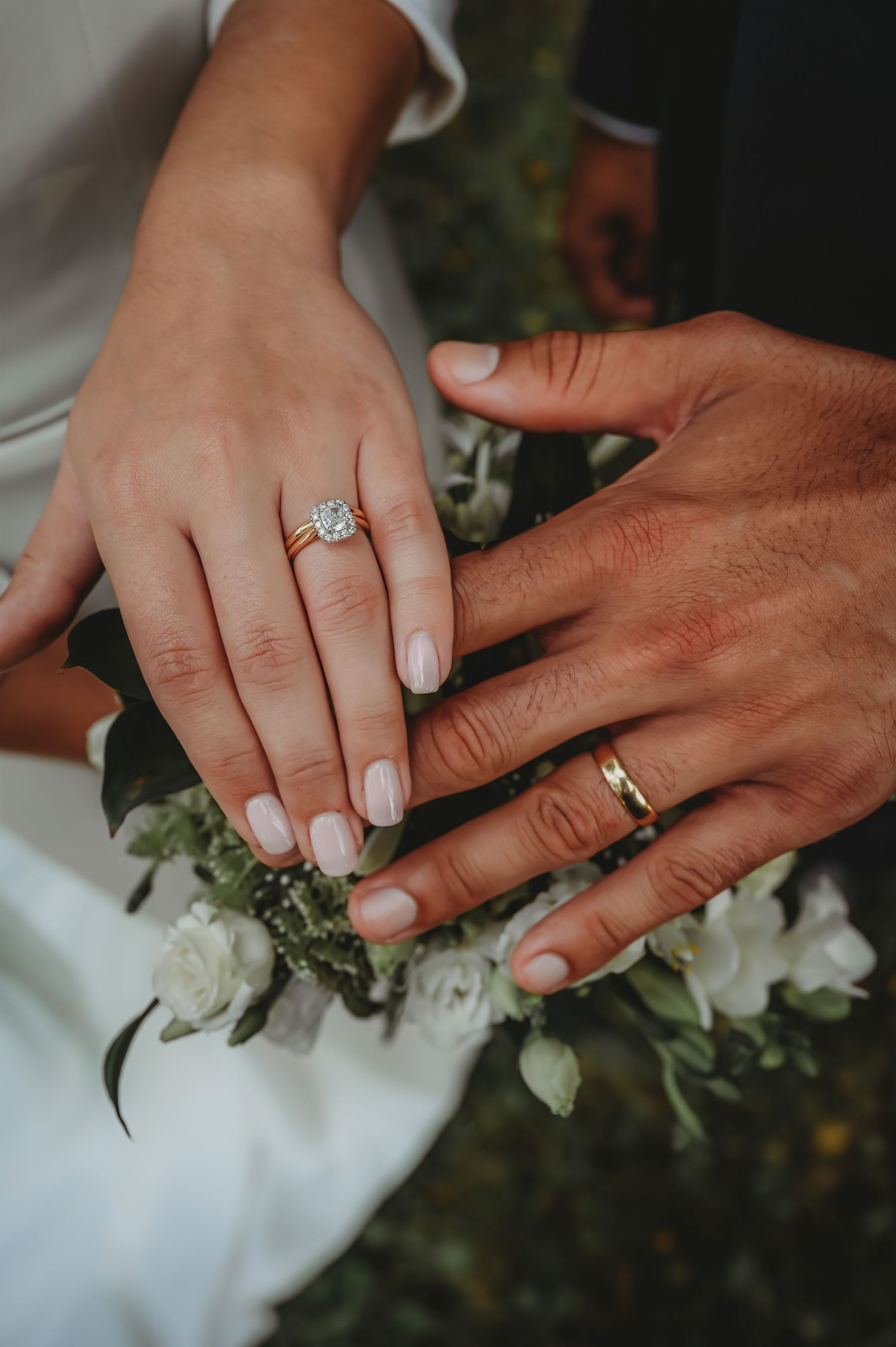 Two hands wearing wedding rings rest together on a white dress with delicate white floral arrangements.