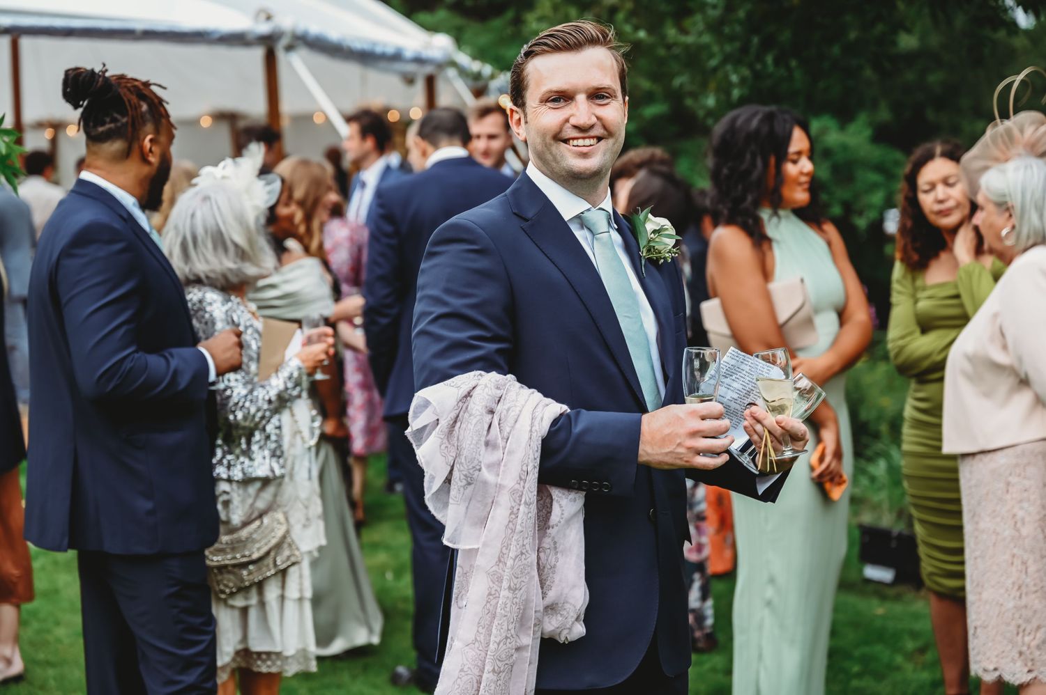 Wedding guests in formal attire mingle during outdoor reception, with person in navy suit holding drink and jacket.