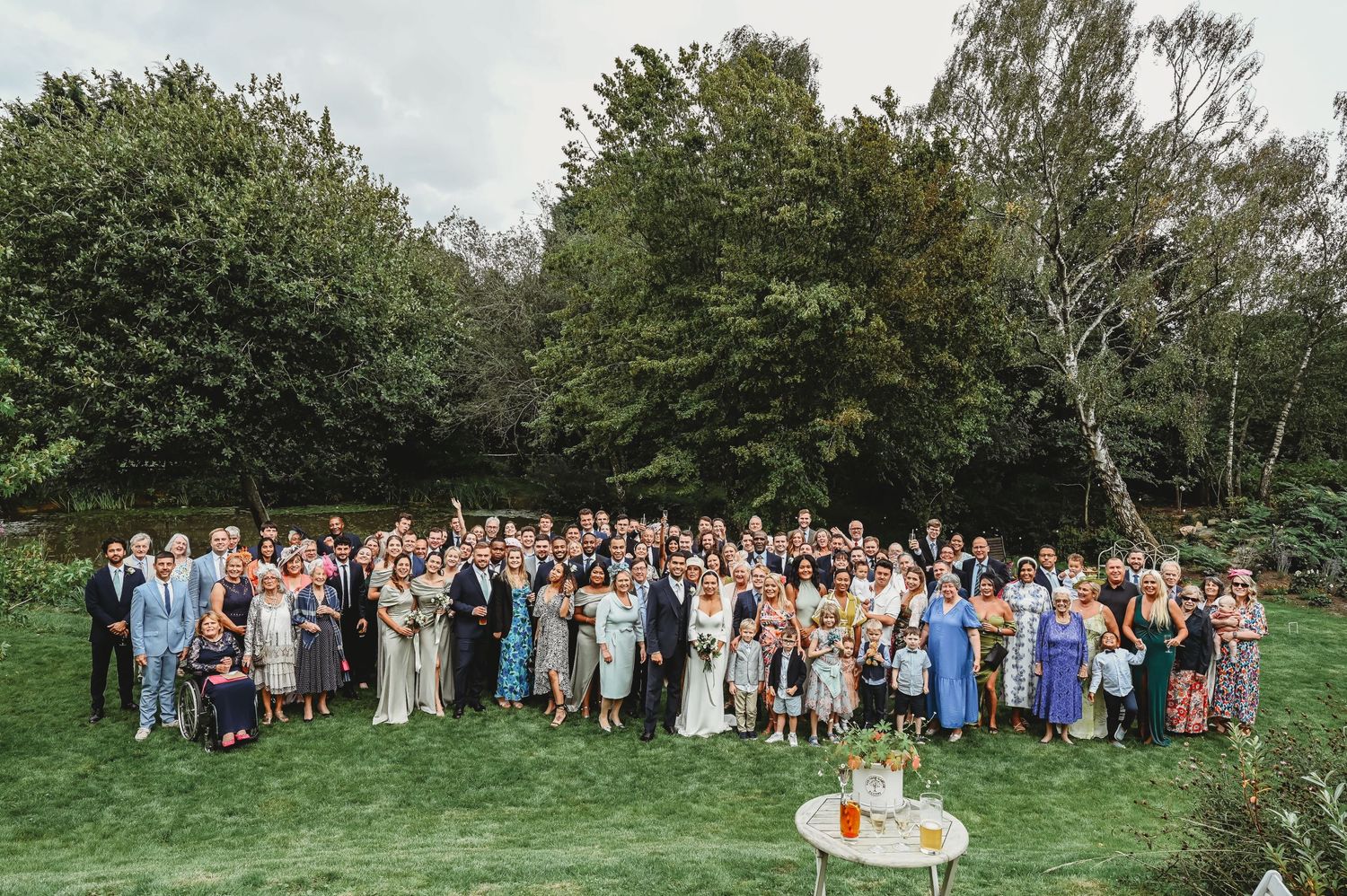 Large outdoor wedding group gathers on green lawn with trees in background during daytime celebration.