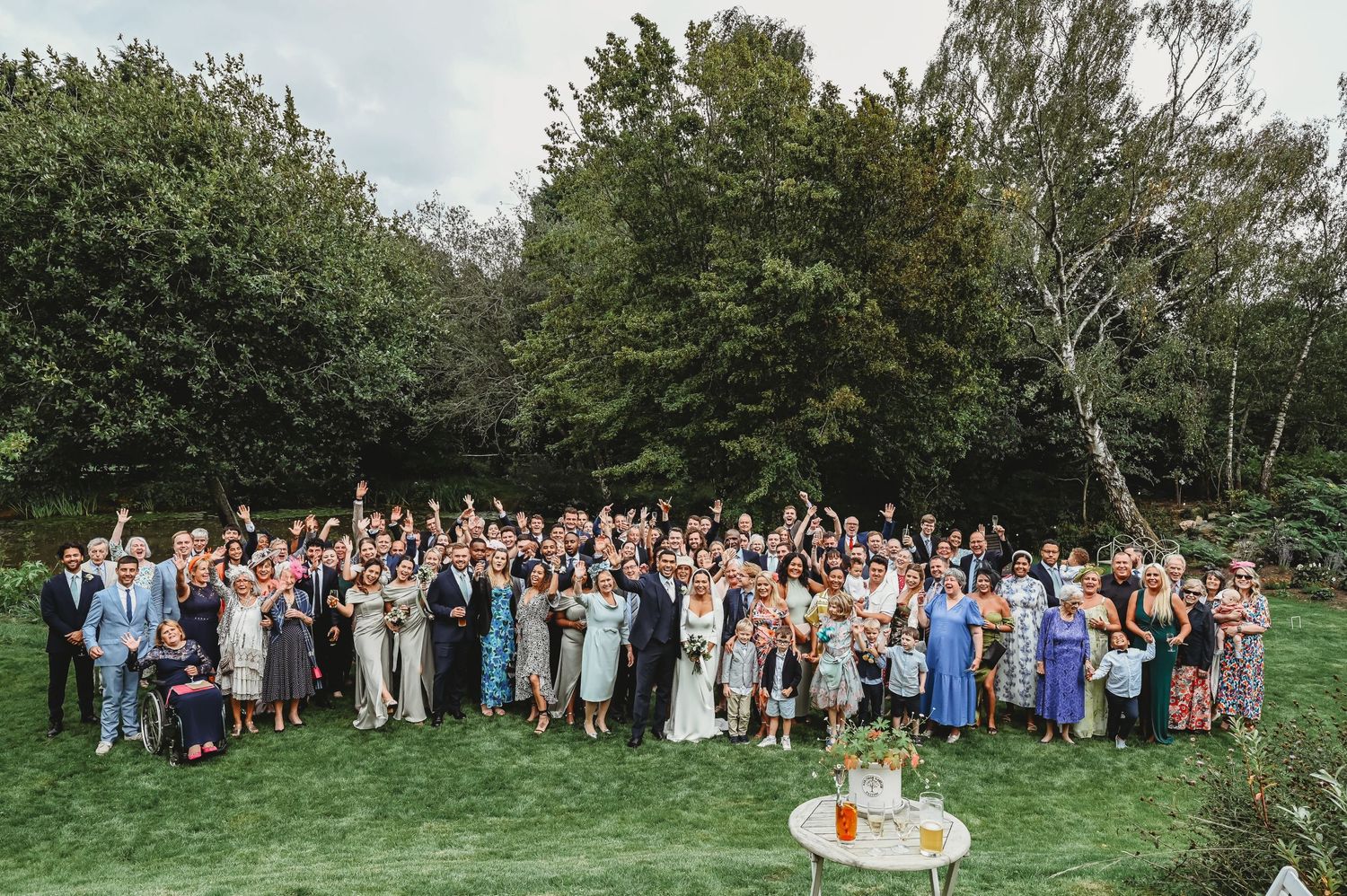 A large group of wedding guests gathered on a green lawn surrounded by trees for an outdoor reception photograph.