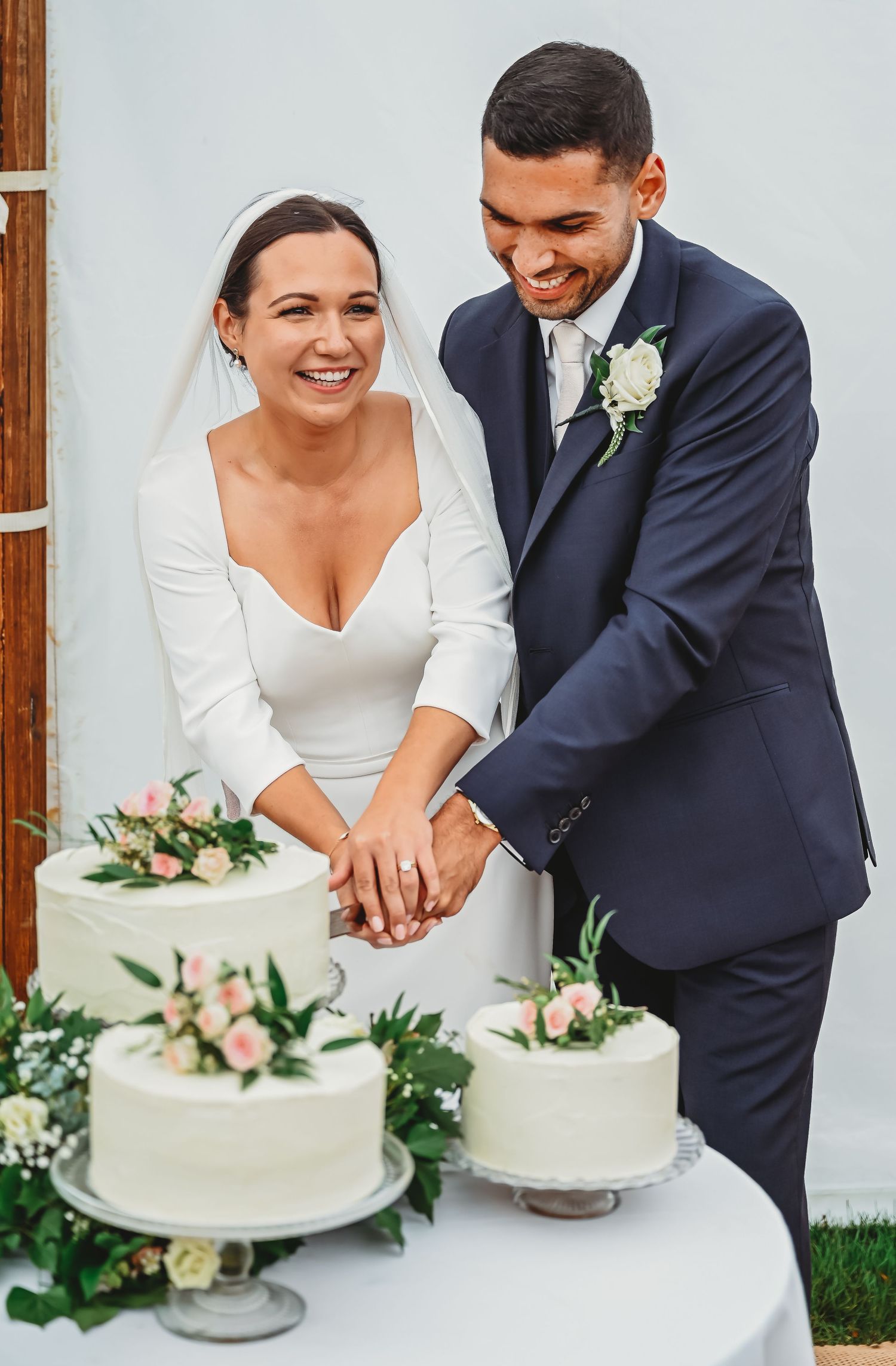 Happy couple cuts their elegant white wedding cake decorated with blush roses and greenery at outdoor reception.
